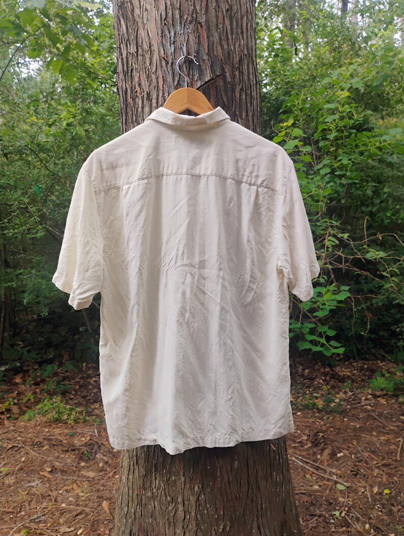 May include: Cream-colored short-sleeve shirt hanging on a wooden hanger. The shirt has a button-down front and a collar. The back of the shirt is visible, showing a yoke detail. The shirt is made of a lightweight fabric.