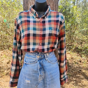 May include: A button-down flannel shirt in a plaid pattern of orange, teal, navy, and tan. The shirt is paired with a light blue denim skirt. The outfit is displayed on a black mannequin.