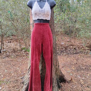 May include: A white crochet halter top and high-waisted, wide-leg, burgundy corduroy pants displayed on a mannequin. The pants have vertical pleats. The outfit is displayed outdoors, leaning against a tree.