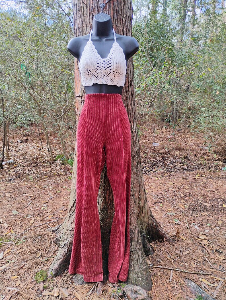 May include: A white crochet halter top and high-waisted, wide-leg, burgundy corduroy pants displayed on a mannequin. The pants have vertical pleats. The outfit is displayed outdoors, leaning against a tree.