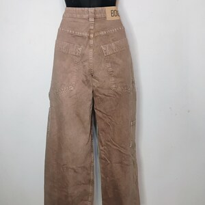 May include: High-waisted brown denim pants with a wide-leg silhouette, featuring large back pockets, a utility-style side pocket, and a tan leather-colored brand patch on the waistband with the visible text BDG printed in black.