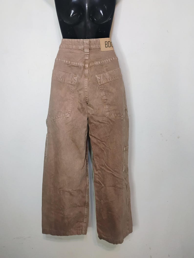 May include: High-waisted brown denim pants with a wide-leg silhouette, featuring large back pockets, a utility-style side pocket, and a tan leather-colored brand patch on the waistband with the visible text BDG printed in black.