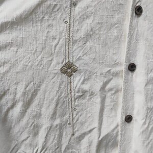 May include: Close-up of a cream-colored button-down shirt. The shirt features a textured fabric and a vertical embroidered design with a geometric pattern. Wooden buttons are visible along the placket.