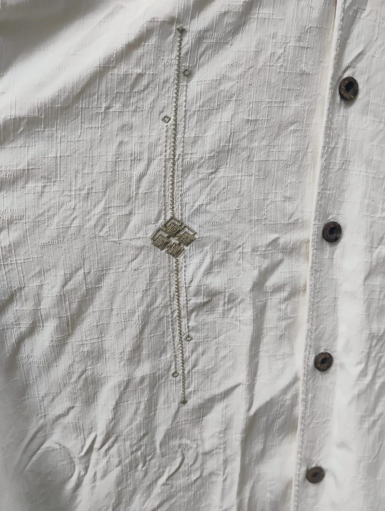 May include: Close-up of a cream-colored button-down shirt. The shirt features a textured fabric and a vertical embroidered design with a geometric pattern. Wooden buttons are visible along the placket.