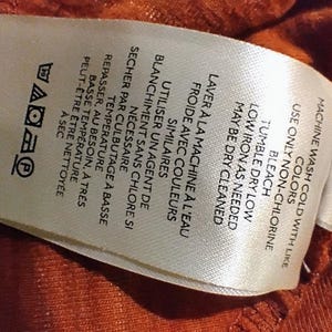May include: A white fabric tag with black text and laundry symbols. The tag includes instructions for machine washing in cold water with like colors, tumble drying on low, and low ironing. The tag also has French text.