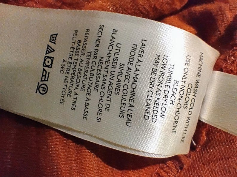 May include: A white fabric tag with black text and laundry symbols. The tag includes instructions for machine washing in cold water with like colors, tumble drying on low, and low ironing. The tag also has French text.