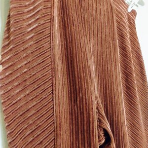 May include: Close-up of brown pleated pants hanging on a white hanger. The pants have a vertical ribbed texture with a diagonal ribbed pattern on the side. The fabric appears to be soft and flowing.