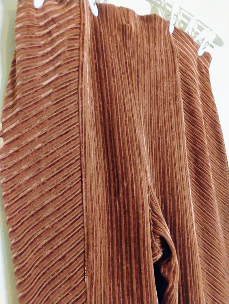 May include: Close-up of brown pleated pants hanging on a white hanger. The pants have a vertical ribbed texture with a diagonal ribbed pattern on the side. The fabric appears to be soft and flowing.