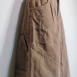 May include: A pair of brown denim pants featuring a high-waisted design with a button fly closure, deep side pockets, and reinforced stitching along the seams. The fabric has a visible twill texture and includes metal rivets at the pocket corners.