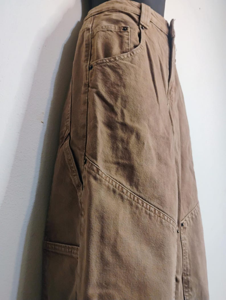 May include: A pair of brown denim pants featuring a high-waisted design with a button fly closure, deep side pockets, and reinforced stitching along the seams. The fabric has a visible twill texture and includes metal rivets at the pocket corners.