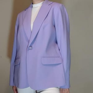 May include: A light purple blazer with a single button closure and two front pockets. The blazer is worn over a white turtleneck. The blazer has long sleeves and a notched lapel.