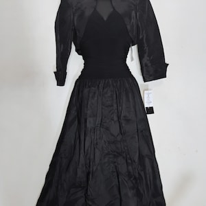 May include: A black vintage dress with a matching bolero jacket. The dress features a fitted bodice, a cinched waist, and a flowing skirt. The bolero has three-quarter sleeves and a high collar. The dress is displayed on a mannequin against a white background.