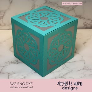 May include: A teal paper cube with intricate cut-out designs on each side. The cube has a gray interior, and the text "INSTANT DOWNLOAD" is visible. The bottom of the cube has the text "SVG PNG DXF instant download" and the designer's name.