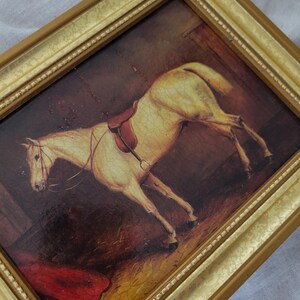 May include: A framed painting of a white horse with a brown saddle in a stable setting. The horse is standing on straw and the background is a dark brown.