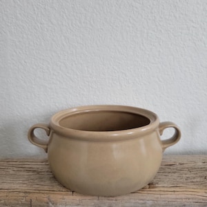 May include: A beige ceramic serving bowl with two handles. The bowl is round and has a slightly flared rim.