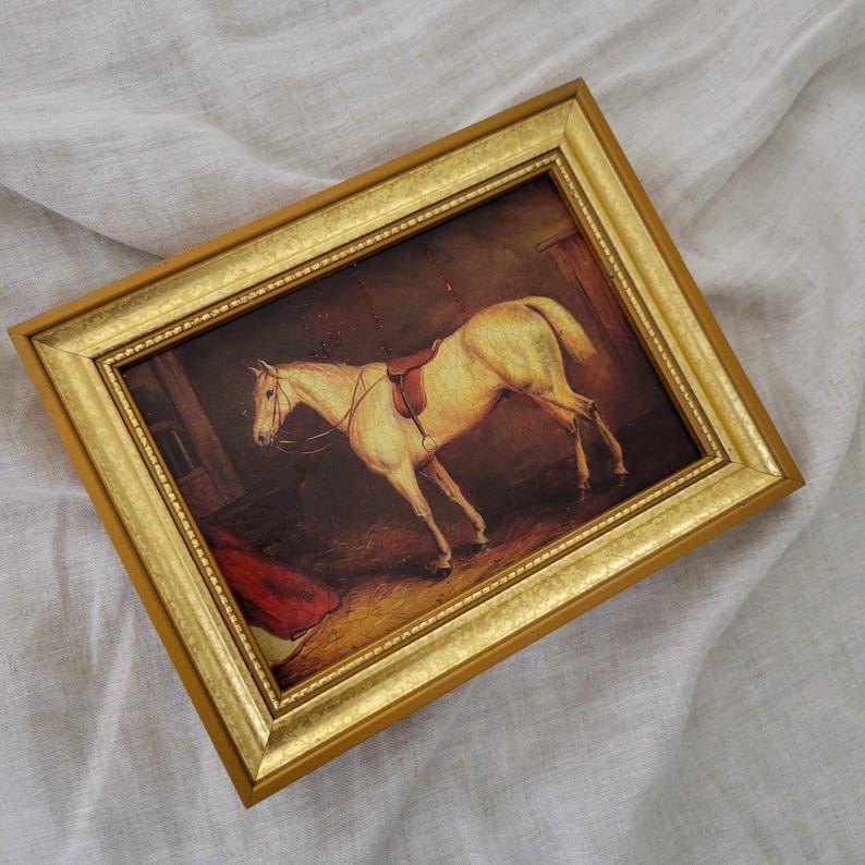 May include: A framed painting of a white horse with a saddle in a stable setting. The horse is standing in a dimly lit room with a red blanket in the background. The frame is gold with a decorative border.