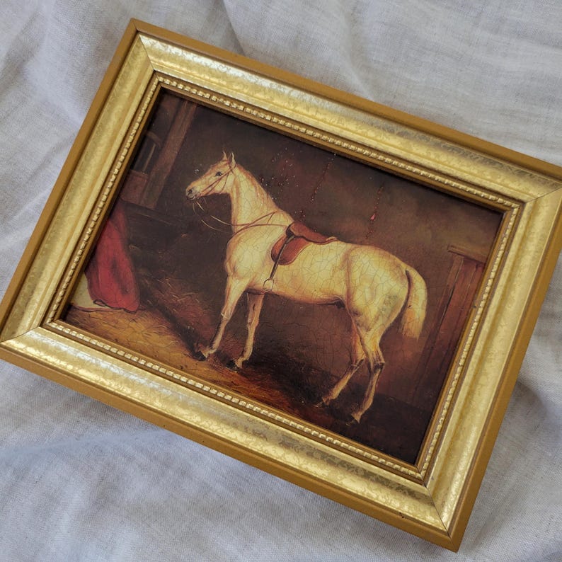 May include: A framed painting of a white horse with a saddle in a stable setting. The horse is standing in a stall with a red blanket hanging in the background. The painting is framed in a gold-colored frame.
