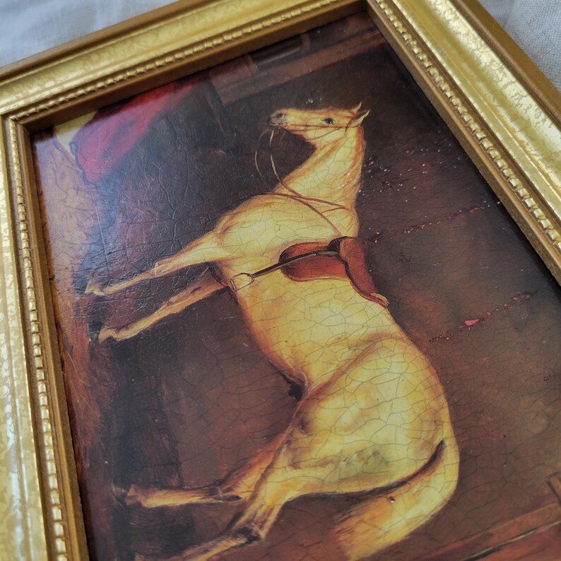 May include: A framed painting of a white horse with a brown saddle, set against a dark brown background. The horse is positioned in a dynamic pose, with its head turned to the side and its legs extended.