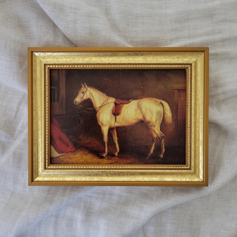 May include: A framed painting of a white horse in a stable. The horse is wearing a saddle and bridle. The painting is in a gold frame.