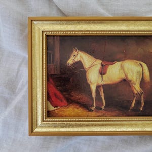 May include: A framed painting of a white horse with a brown saddle in a stable setting. The horse is standing in a dimly lit barn with a red blanket in the background.