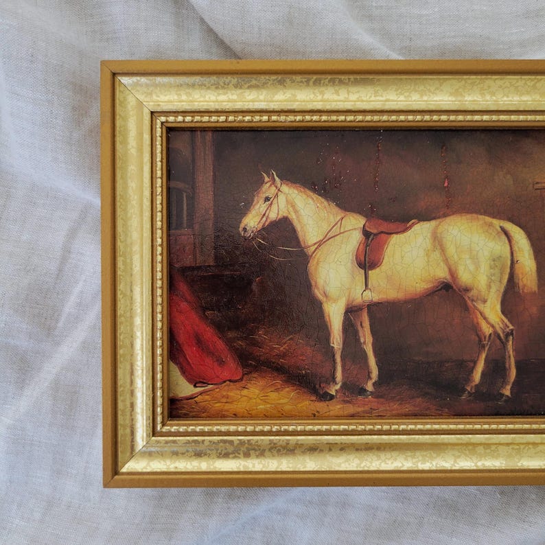 May include: A framed painting of a white horse with a brown saddle in a stable setting. The horse is standing in a dimly lit barn with a red blanket in the background.