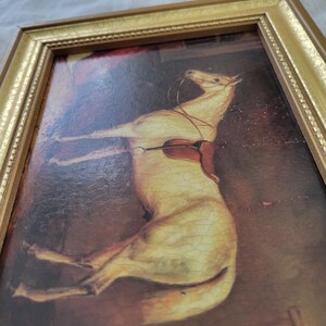 May include: A framed painting of a white horse with a saddle, set against a dark brown background. The horse is depicted in a dynamic pose, with its head tilted back and its legs outstretched.
