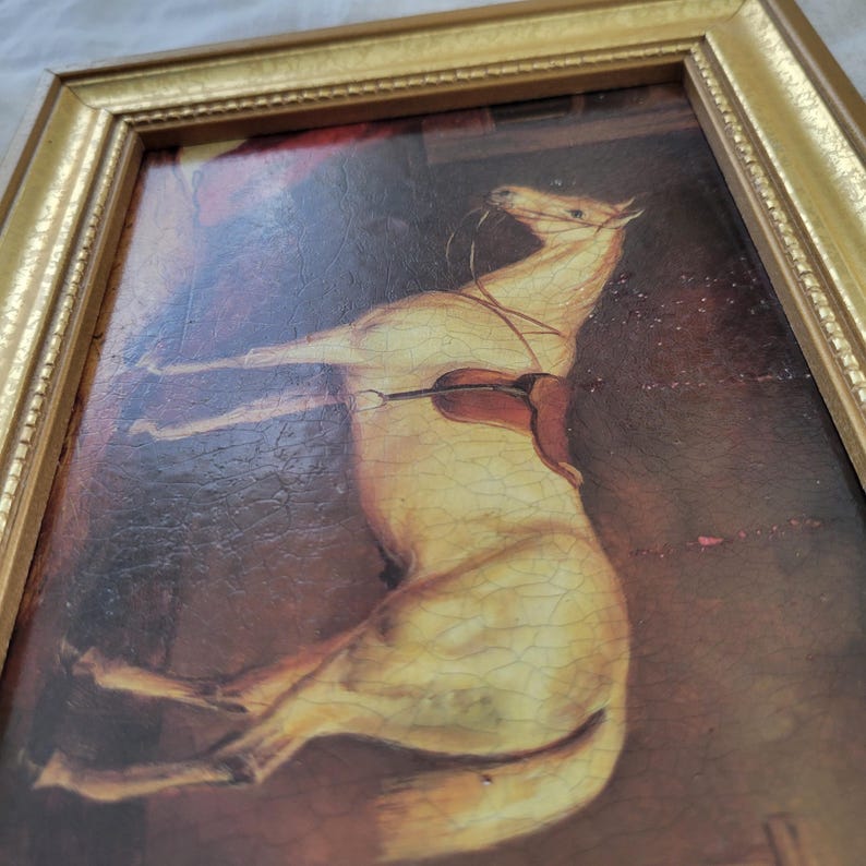 May include: A framed painting of a white horse with a saddle, set against a dark brown background. The horse is depicted in a dynamic pose, with its head tilted back and its legs outstretched.
