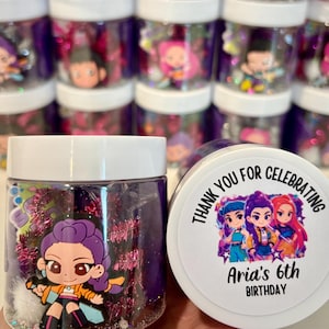 Op de afbeelding: Transparante plastic potten gevuld met paarse glitter en karakterbeeldjes. De potten hebben witte deksels. Eén pot heeft een label met de tekst "Bedankt voor het vieren van Aria's 6e verjaardag".