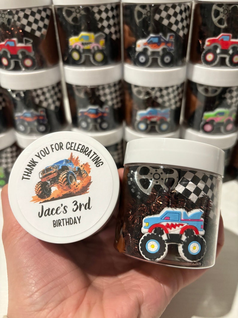 Op de afbeelding: Een ronde witte bedankbox met een monstertruckontwerp en de tekst "Bedankt voor het vieren van Jace's 3e verjaardag". De doos is gevuld met bruin en zwart slijm en een blauw monstertruckspeelgoed.