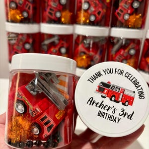 May include: Red and black toy fire engine in a clear plastic jar with a white lid. The lid has a white sticker with black text that says "Thank you for celebrating Archer's 3rd Birthday".