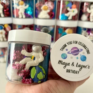 Puede incluir: Un tarro de plástico transparente lleno de purpurina rosa y azul, una pequeña figura de astronauta y un planeta azul y verde. El tarro tiene una tapa blanca con una pegatina que dice "Gracias por celebrar el cumpleaños de Maya & Layne".