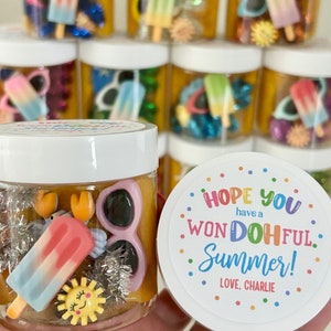 Peut inclure: Un pot transparent rempli de pâte à modeler jaune et de divers jouets sur le thème de l'été, dont une sucette glacée, des lunettes de soleil et un soleil. Le pot a un couvercle blanc avec un autocollant coloré qui dit "J'espère que vous avez un été WONDOhFUL ! AVEC AMOUR, CHARLIE."