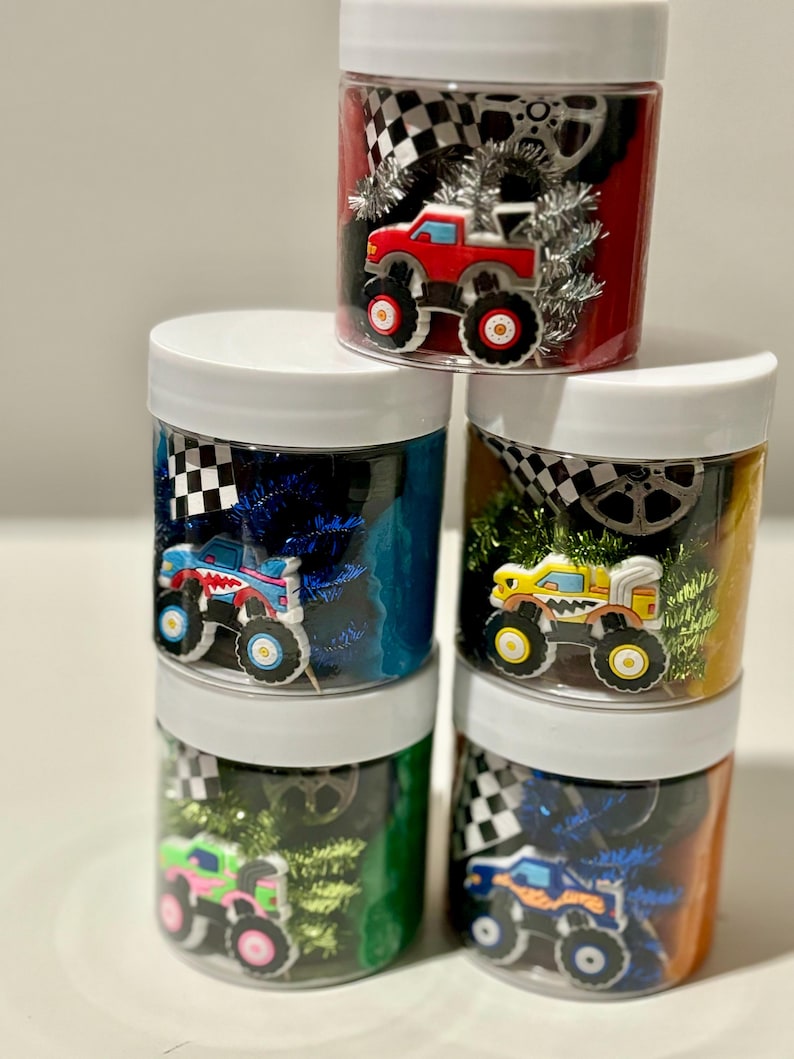 Op de afbeelding: Vier doorzichtige potten gevuld met kleurrijk klei, monstertruck speelgoed en andere kleine speelgoedjes. De potten hebben witte deksels.