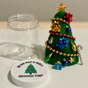 Puede incluir: Un tarro blanco con un diseño de árbol de Navidad verde en la tapa. La tapa dice "¿Quieres construir un árbol de Navidad?" El tarro está vacío y al lado hay un árbol de Navidad de plastilina verde decorado con cuentas doradas, lazos rojos y azules y un bastón de caramelo.