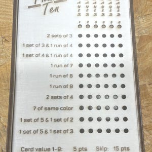 Peut inclure: Tableau de score en bois pour le jeu Phase 10, avec le titre du jeu en haut. Le tableau comporte une série de trous et du texte indiquant les exigences de score du jeu. Le texte comprend les valeurs de points pour les valeurs de cartes, le skip et les jokers.