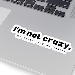 May include: A white sticker with the text "I'm not crazy, my mother had me tested" in black, placed on a laptop. The sticker has a black border and is angled slightly.