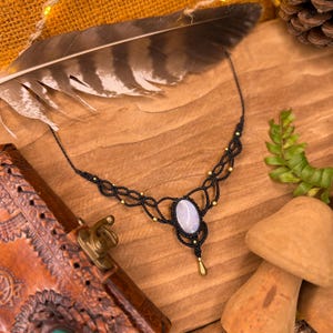 May include: A black macrame necklace featuring a light blue oval stone pendant and gold-coloured beads. The necklace is presented on a wooden surface, alongside a feather, a leather journal, and wooden mushroom ornaments.