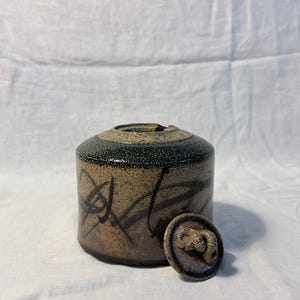 May include: A cylindrical ceramic jar with a speckled tan and dark brown glaze featuring black abstract brushstroke markings. The jar has a matching circular lid that rests beside it, topped with a small, coiled, sculptural ceramic snail figure.