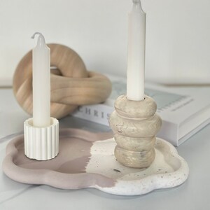 May include: Two white candles in a cloud-shaped tray with a speckled white and light brown glaze. The tray has a light brown and white paint design. One candle is in a white fluted holder, the other is in a light brown holder with a swirl design.