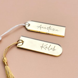 May include: Two gold-colored bookmarks with tassels. The bookmark on the left has a white tassel and the text "Anastasia" engraved on it. The bookmark on the right has a gold tassel and the text "Kaleb" engraved on it.
