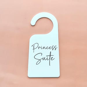 May include: A light blue door hanger with a curved top and the words "Princess Suite" in elegant script. The background is a solid peach color.