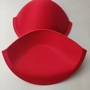 Könnte beinhalten: Zwei rote BH-Schalen aus Schaumstoff. Die Schalen sind halbkreisförmig und haben eine glatte, nahtlose Oberfläche.