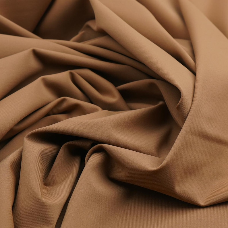 May include: Close-up of a tan fabric with soft folds and a smooth texture. The fabric is a solid color, with subtle variations in tone due to the folds. The material appears to be a medium-weight fabric, suitable for clothing or home decor.