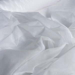 May include: A close-up shot of white tulle fabric, both rolled and draped. The fabric has a fine mesh texture and appears soft and lightweight. The folds and layers create shadows, highlighting the fabric's delicate nature.