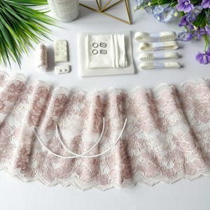 May include: A collection of lingerie-making supplies, including a wide, floral lace fabric in white and mauve, white elastic, bra hooks, rings, and a spool of thread. The items are arranged on a white surface, with decorative flowers and greenery.