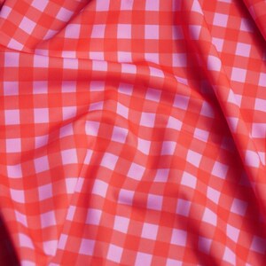 May include: A fabric featuring a vibrant red and light pink gingham check pattern. The material is draped to create soft folds and shadows, highlighting the consistent square grid design across the surface of the textile in a repeating pattern.