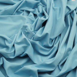 May include: A close-up shot of a light blue fabric with soft folds and a smooth texture. The fabric is draped, creating shadows and highlights that emphasize its fluidity. The color is a muted, pastel shade of blue.