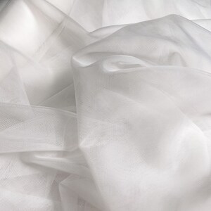 May include: White tulle fabric with a soft, delicate texture. The fabric is sheer and has a fine mesh weave.