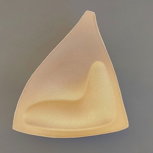 Puede incluir: Una copa de sujetador de espuma beige con una superficie lisa y ligeramente texturizada. La copa tiene forma de triángulo con un fondo redondeado.