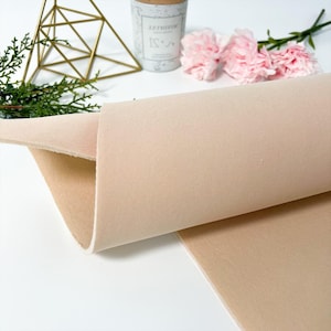 Puede incluir: Un rollo de tela beige, con una superficie suave y texturizada, está parcialmente desenrollado, revelando su grosor. La tela se encuentra sobre un fondo blanco, con elementos decorativos que incluyen una forma geométrica dorada, una pequeña planta y flores rosas.